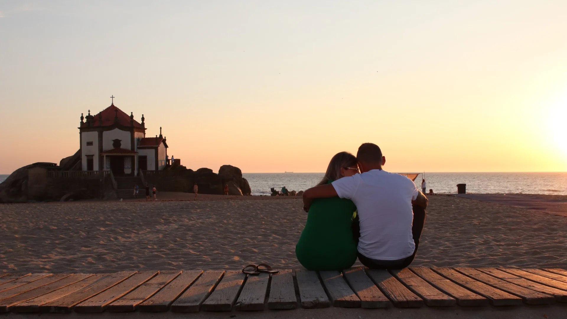 Casal sentado de costas, abraçado, assistindo ao pôr do sol na praia, com uma capela ao fundo, simbolizando um momento de paz e romantismo junto ao mar.