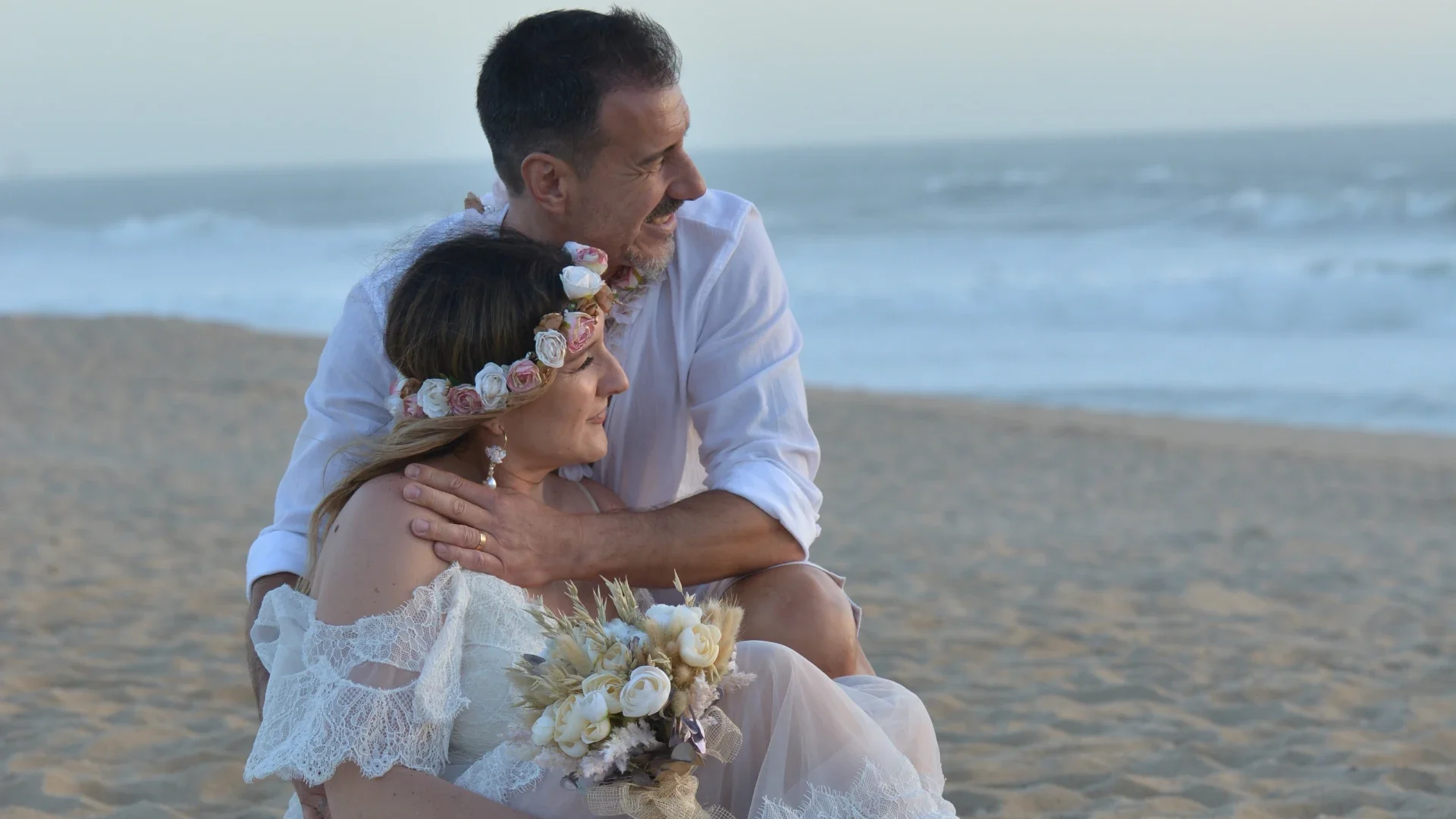 Casal sentado na areia da praia, com o homem abraçando carinhosamente a mulher que segura um buquê de flores, ambos desfrutando de um momento de ternura ao lado do mar.