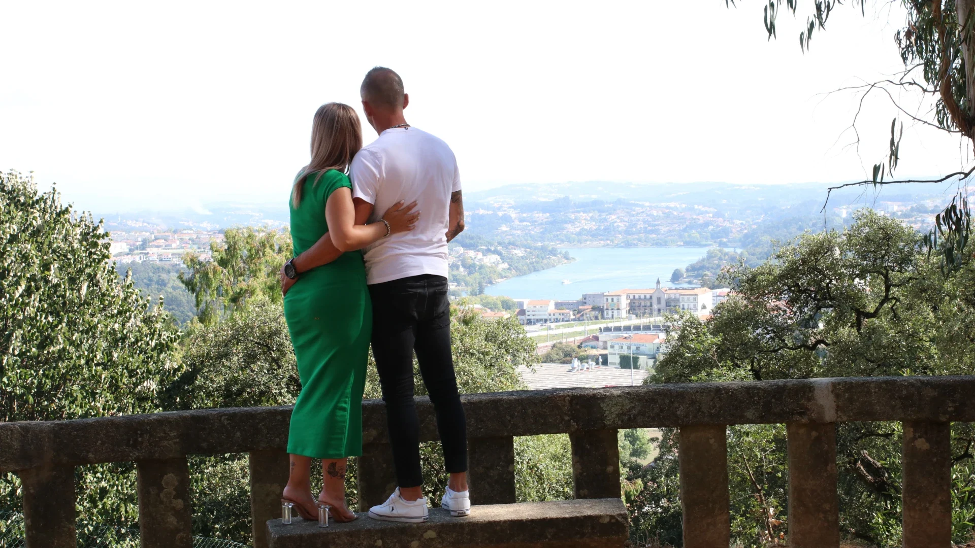 Casal de costas para a câmara, admirando uma vista panorâmica de uma cidade com um rio ao fundo, num momento de tranquilidade e conexão com a natureza.
