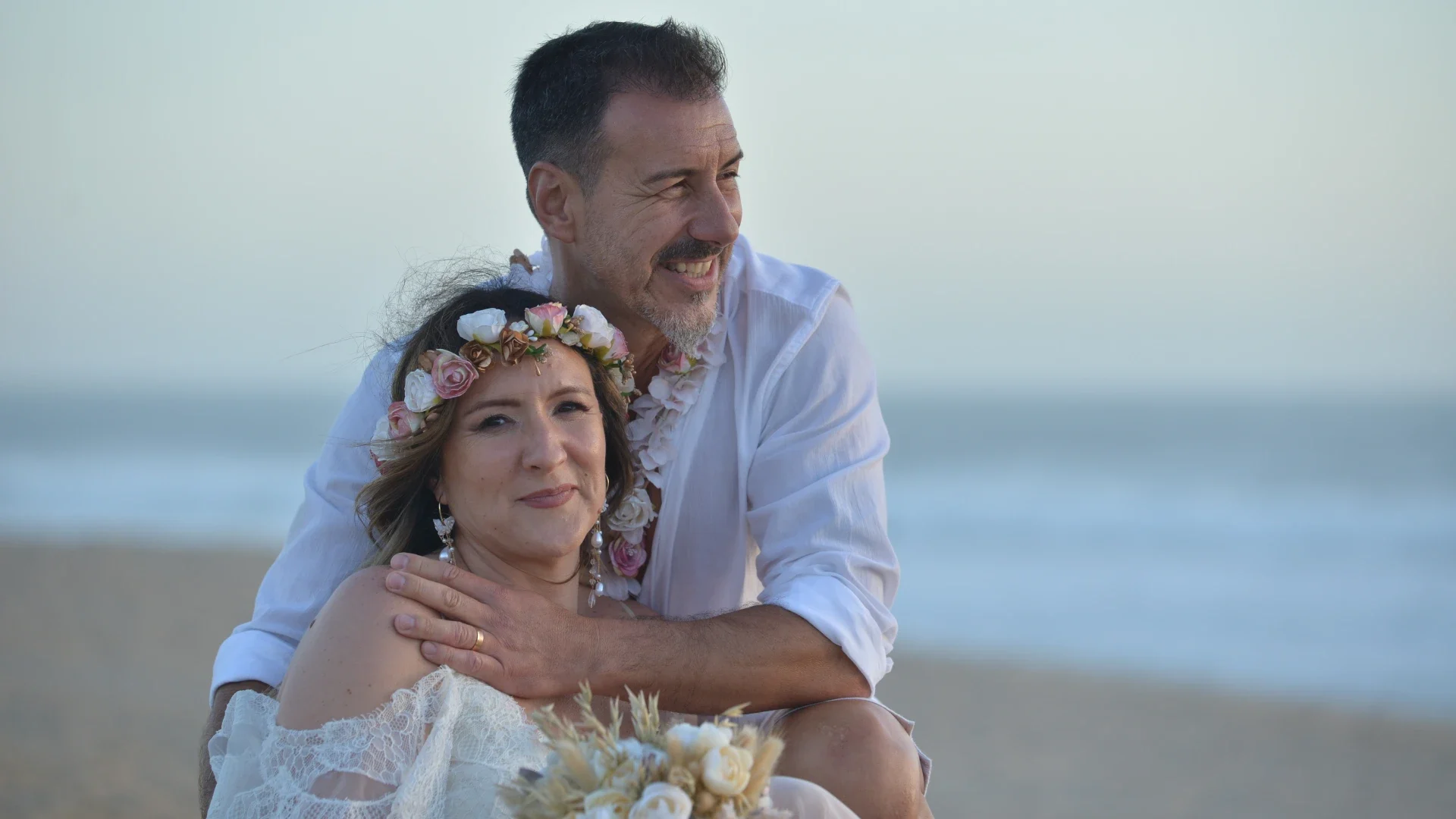 Casal sentado na praia, com o homem abraçando carinhosamente a mulher que usa uma coroa de flores, ambos sorrindo e apreciando um momento especial junto ao mar.