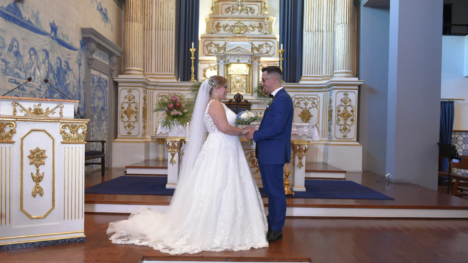 Casal trocando alianças em cerimónia de casamento numa igreja decorada com detalhes dourados e azulejos azuis.