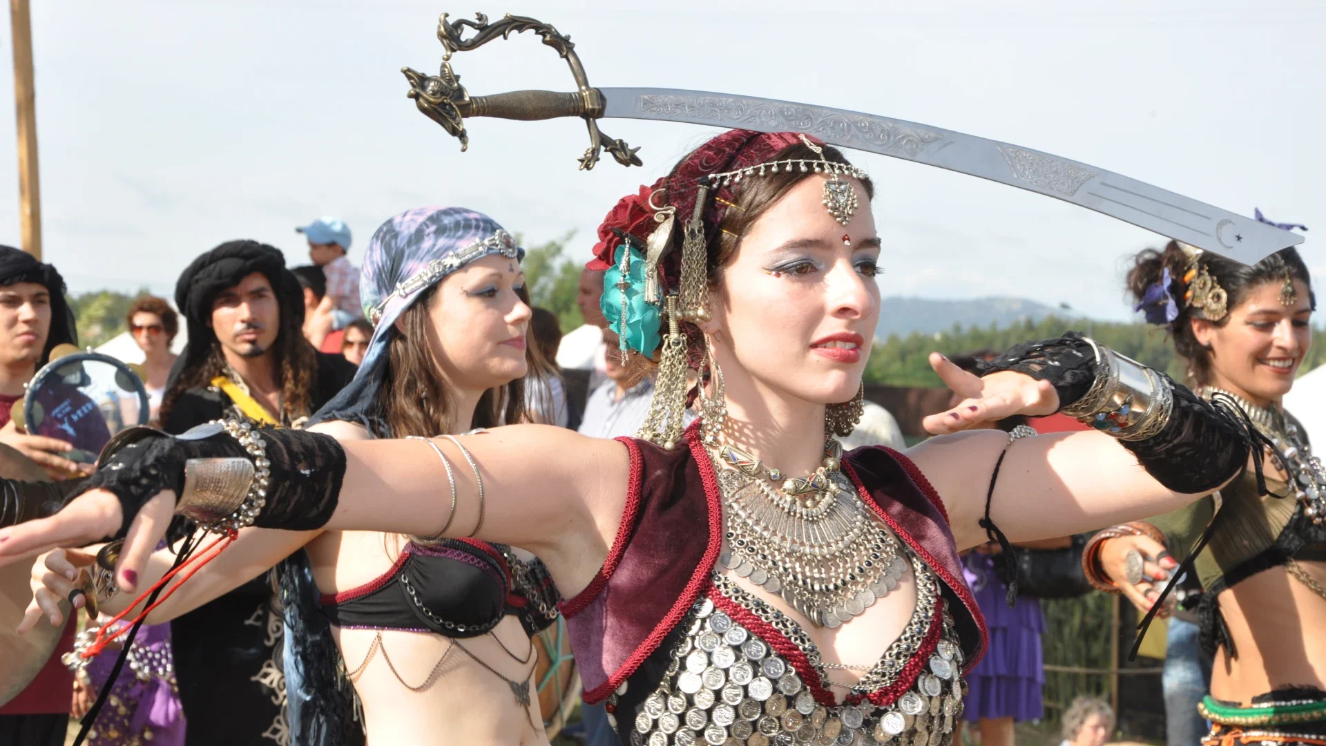 Dançarina em trajes tribais exibindo uma espada equilibrada na cabeça durante uma performance num evento medieval, cercada por outros participantes vestidos em roupas tradicionais, criando uma cena vibrante e culturalmente rica.