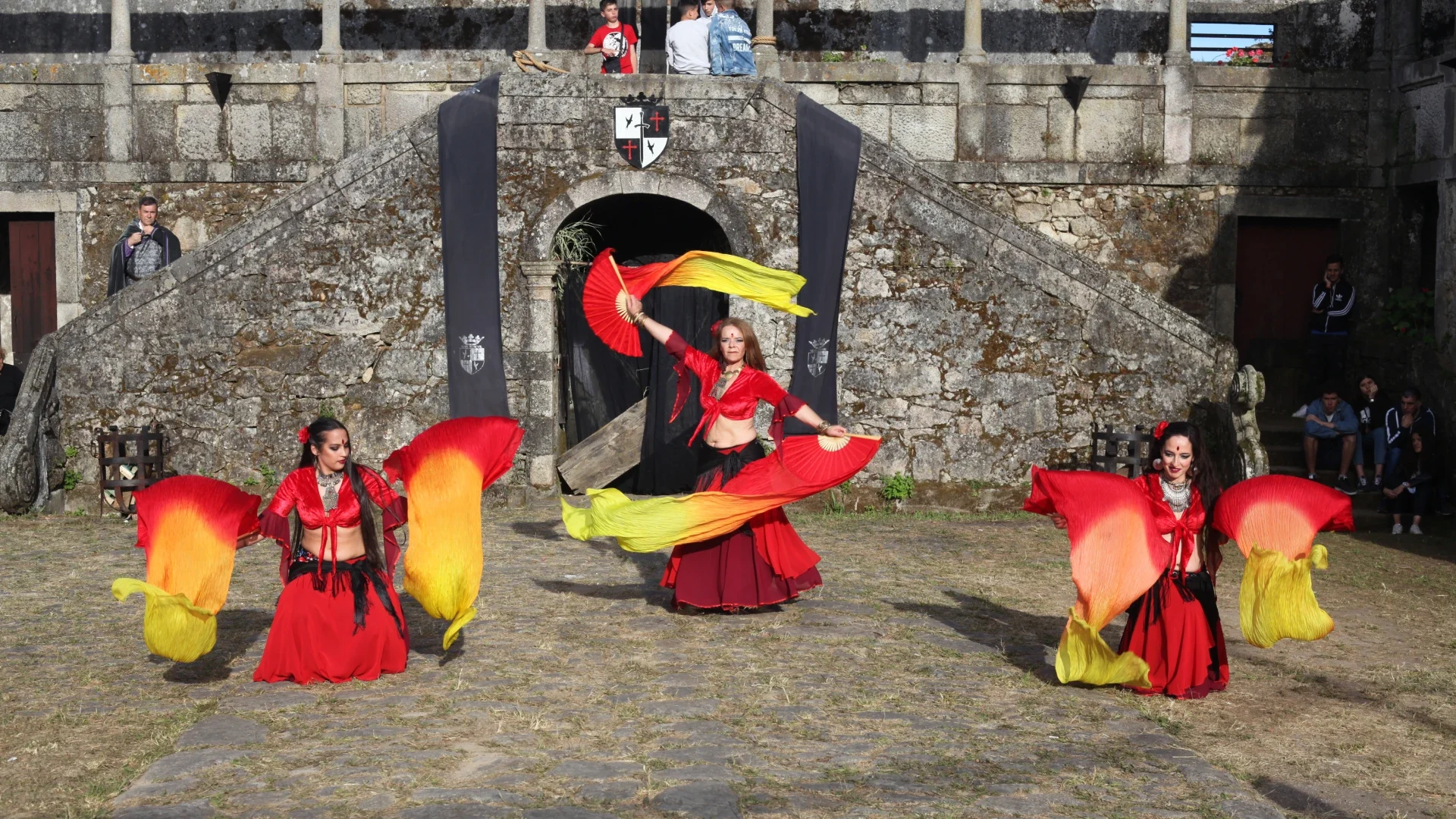 Três dançarinas vestidas em trajes vibrantes, com lenços coloridos, realizando uma apresentação de dança tribal num evento medieval ao ar livre, em frente a uma fortificação de pedra, criando um espetáculo de cor e movimento.