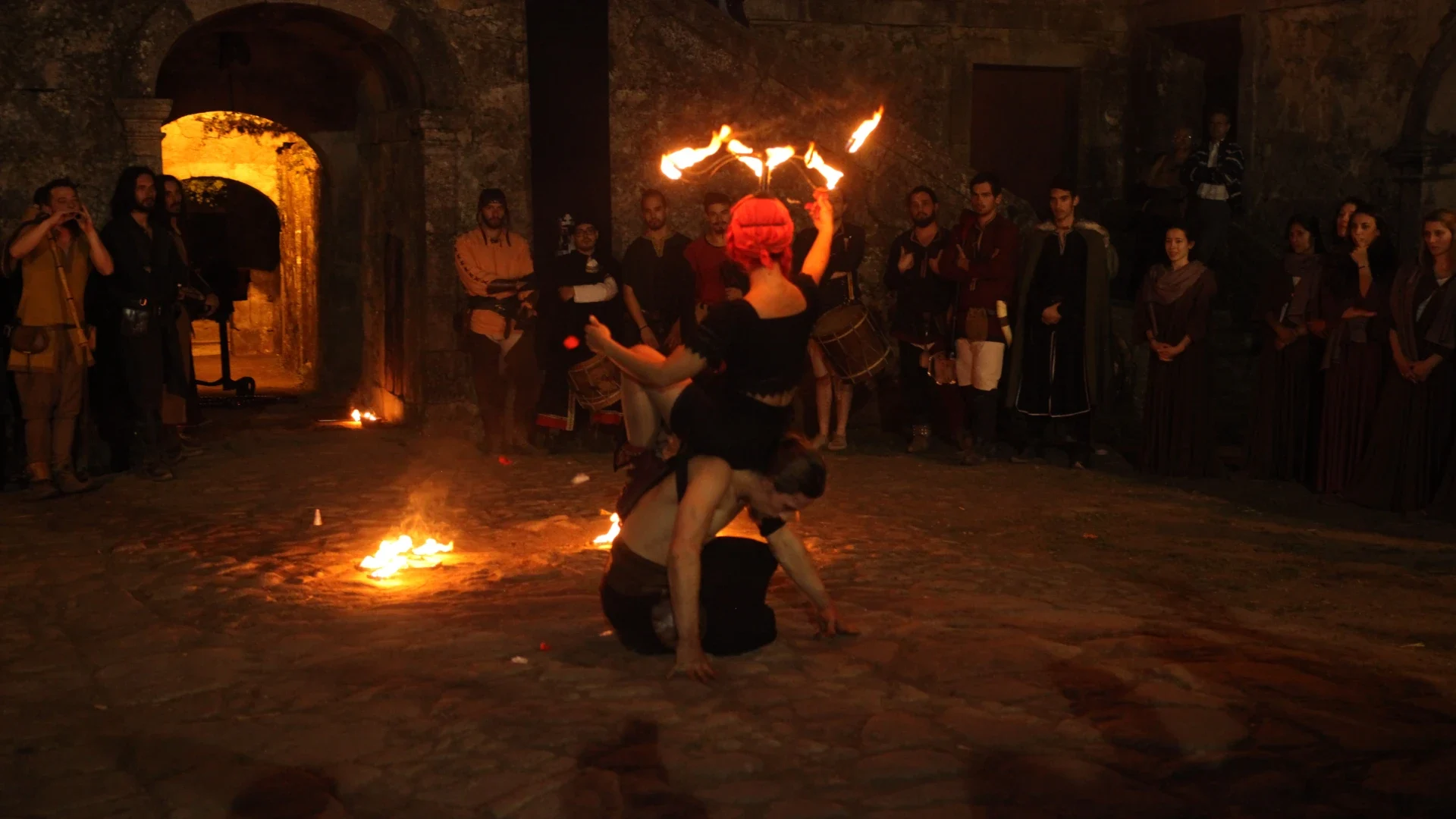 Artistas realizando um espetáculo de malabarismo com fogo durante um evento medieval noturno, cercados por espetadores em trajes históricos, criando uma atmosfera intensa e cativante.