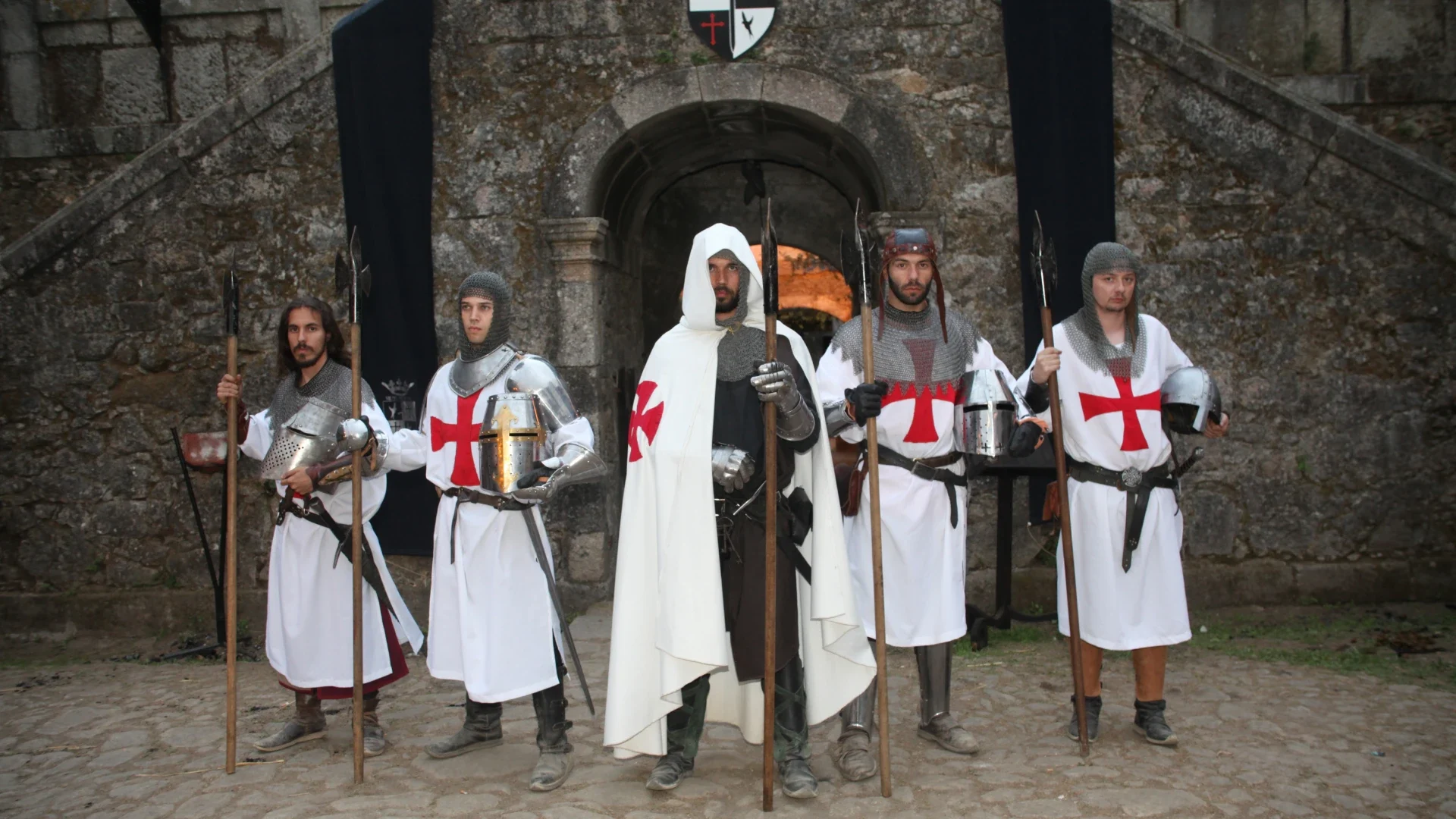 Cinco homens vestidos como Cavaleiros Templários, com armaduras e vestes brancas com cruzes vermelhas, posando em frente a uma fortificação de pedra durante uma recriação histórica medieval.