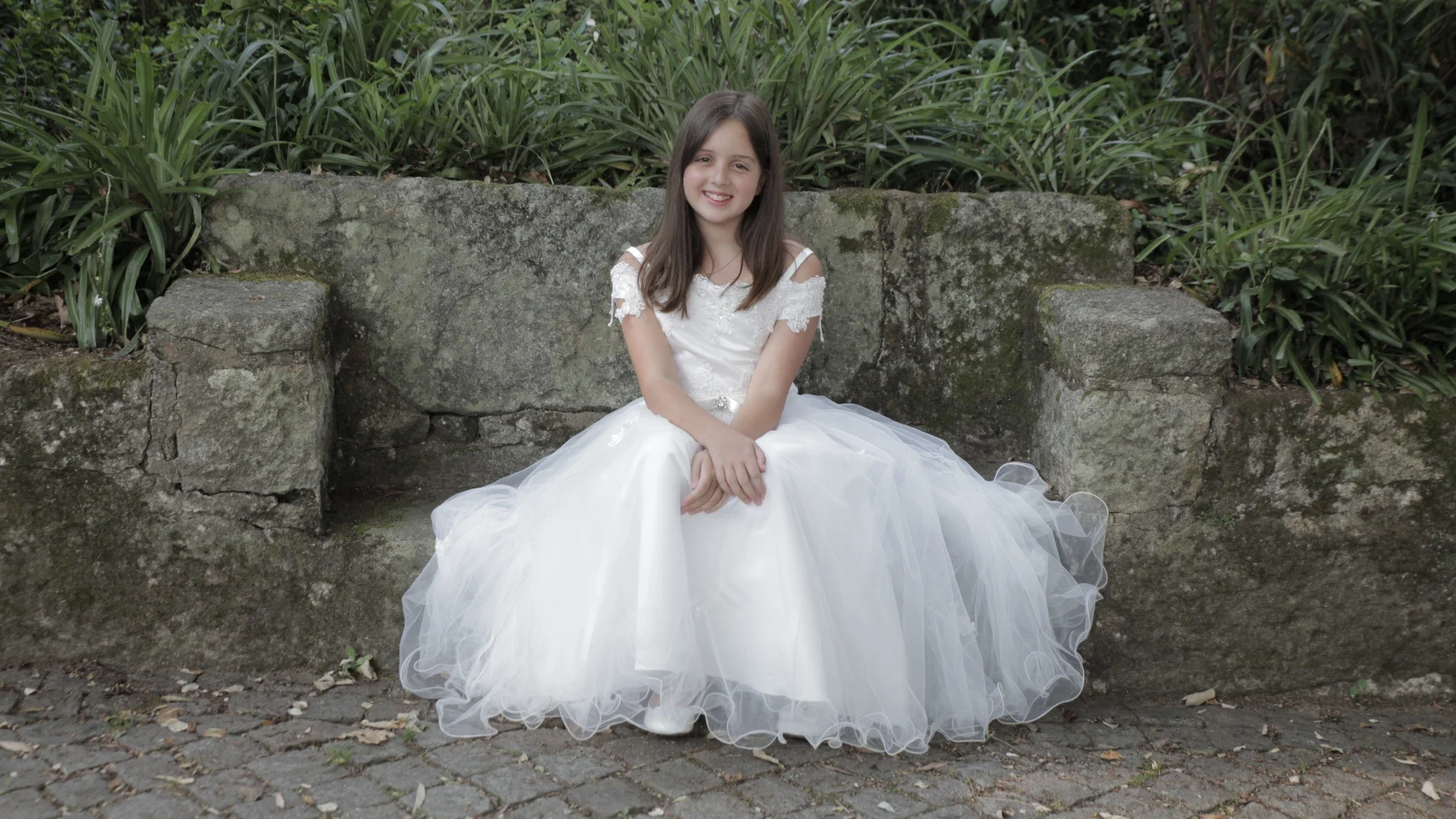 Menina sentada num banco de pedra, usando um vestido branco elegante para a sua Primeira Comunhão, sorrindo serenamente num ambiente natural.
