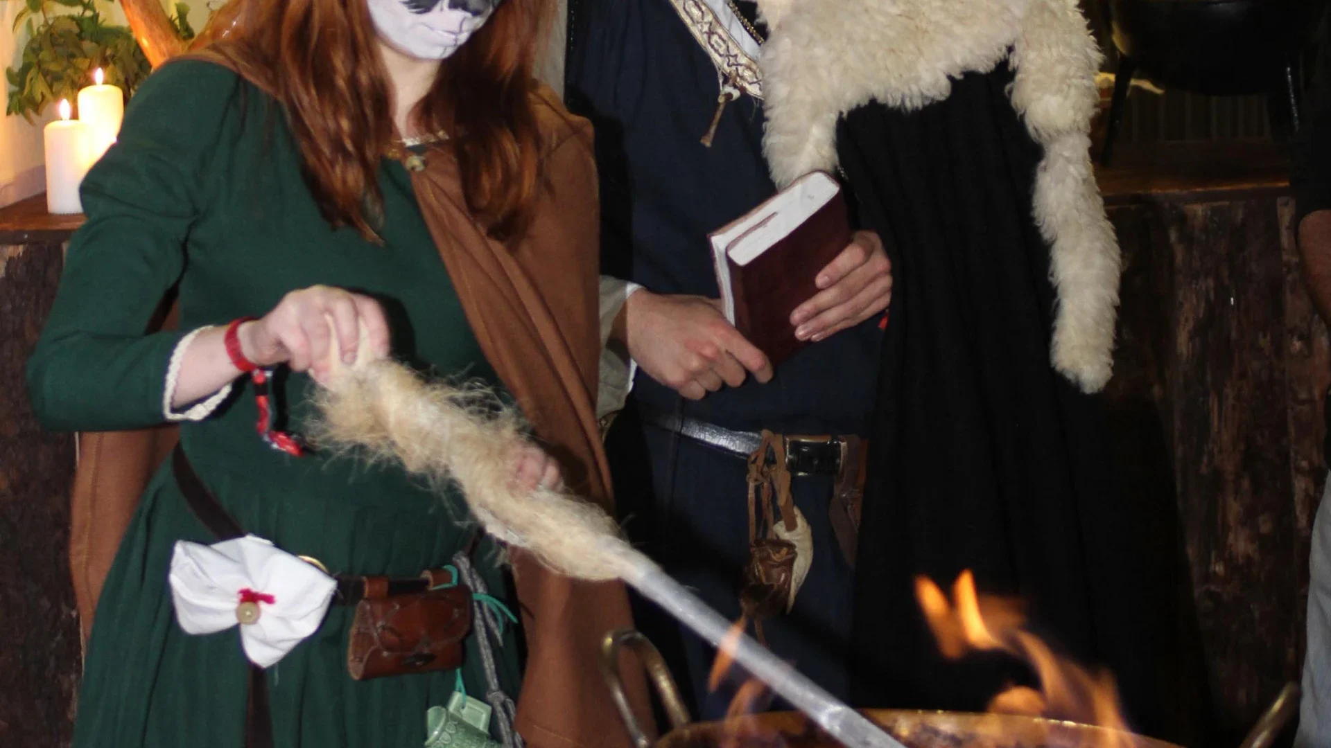 Pessoa vestida com traje medieval verde, usando uma máscara pintada no rosto, demonstrando uma atividade tradicional ao lado de um fogo durante um evento histórico, com outra pessoa ao fundo segurando um livro.